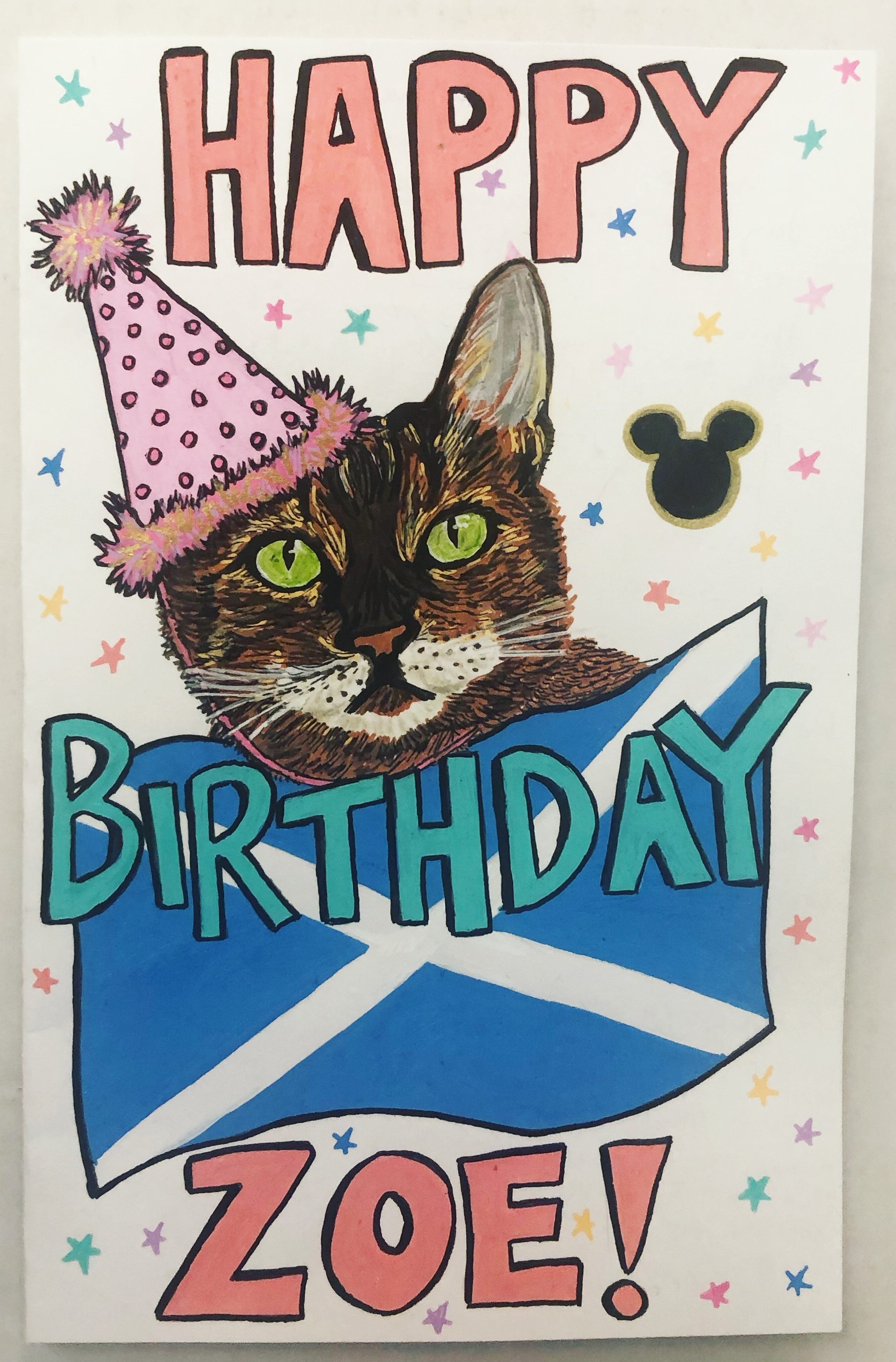 A birthday card I made for a friend with a drawing of her cat wearing a party hat behind a Scottish flag, I created the card with acrylic markers and ink.